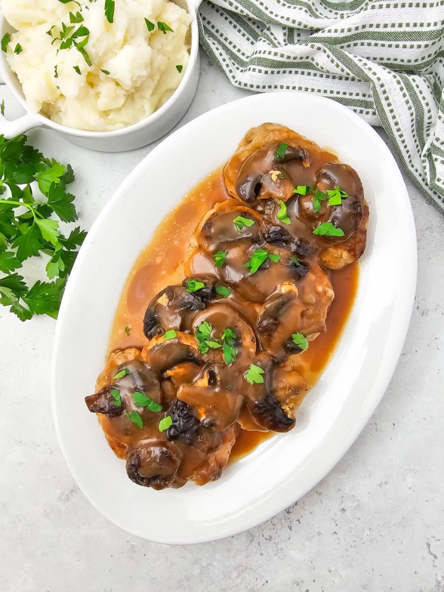 Overhead view of pork chops and mushrooms covered in brown gravy on a white platter, with a white bowl of mashed potatoes and fresh parsley sprigs in the background.