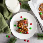 An overhead flat lay featuring a white plate with stacked Reindeer Chocolate Bark, a glass of milk, a green napkin, and scattered red and green M&M candies.