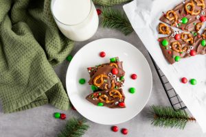 An overhead flat lay featuring a white plate with stacked Reindeer Chocolate Bark, a glass of milk, a green napkin, and scattered red and green M&M candies.