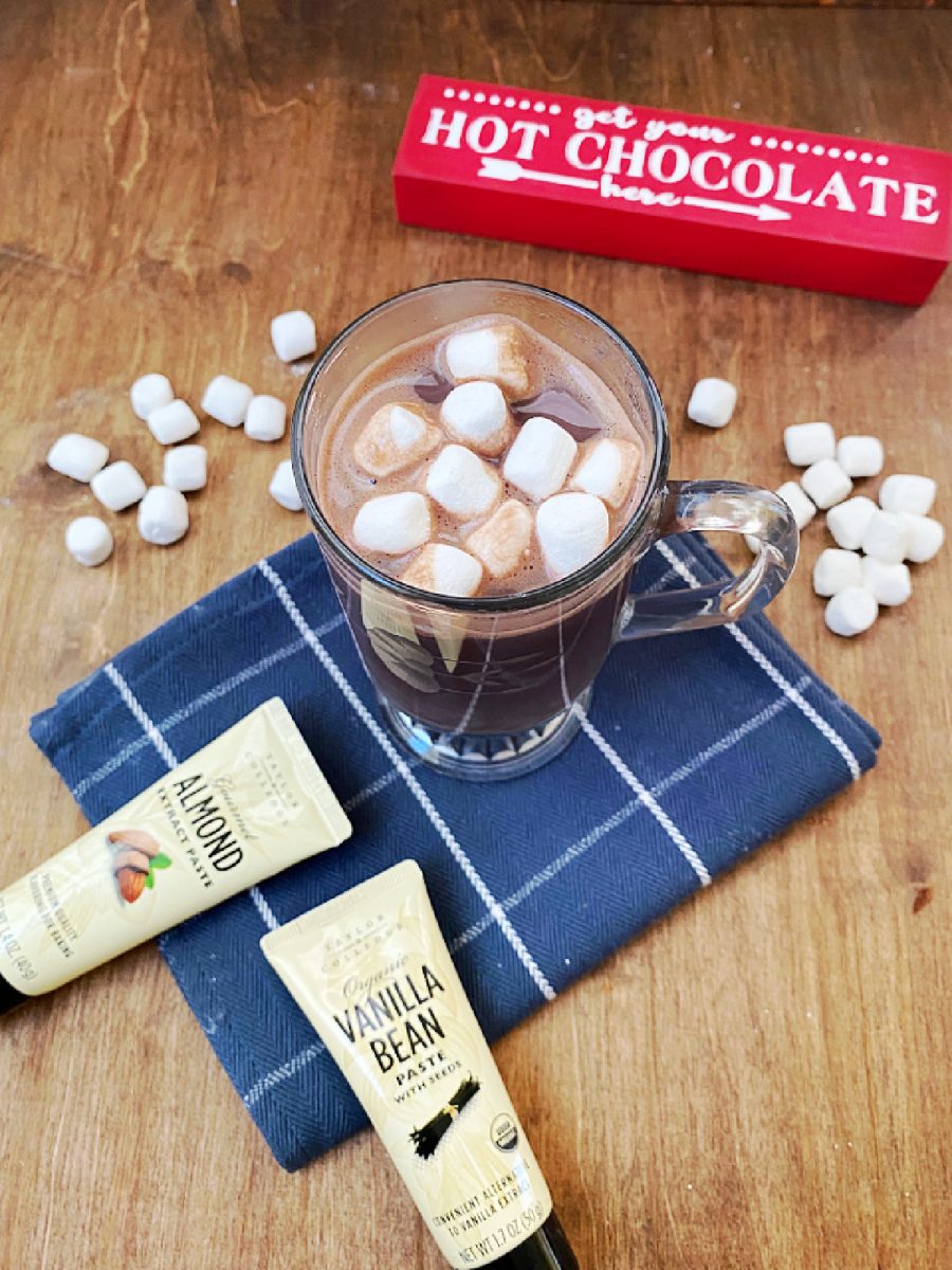 Overhead view of the finished gourmet hot chocolate served in a clear mug, topped with marshmallows, next to the Taylor & Colledge vanilla and almond flavor pastes.