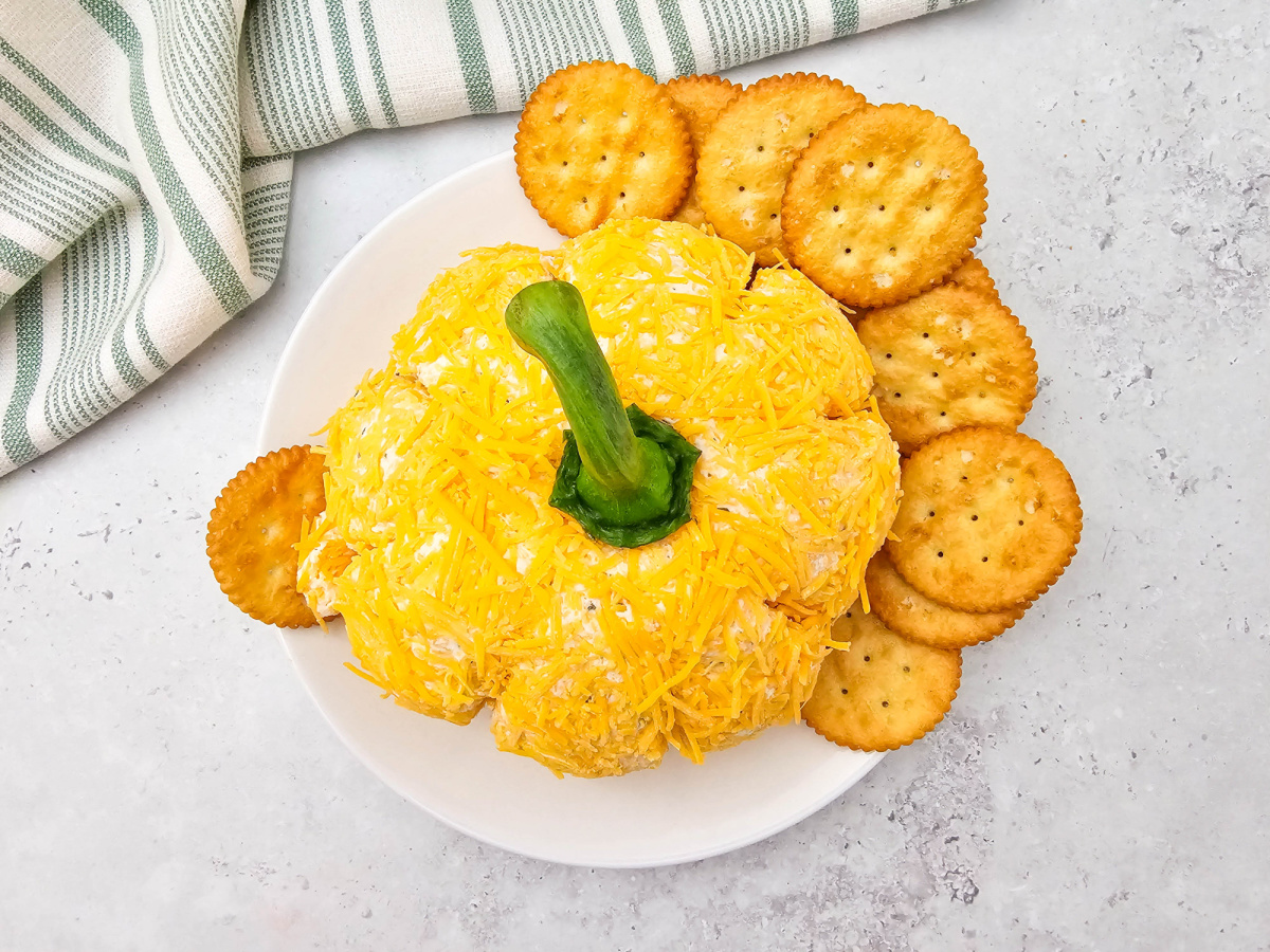 An overhead shot of the Pumpkin Cheese Ball presented on a white plate. The cheese ball is coated in shredded cheddar and shaped like a pumpkin with a green bell pepper stem. It is surrounded by numerous round, butter crackers.