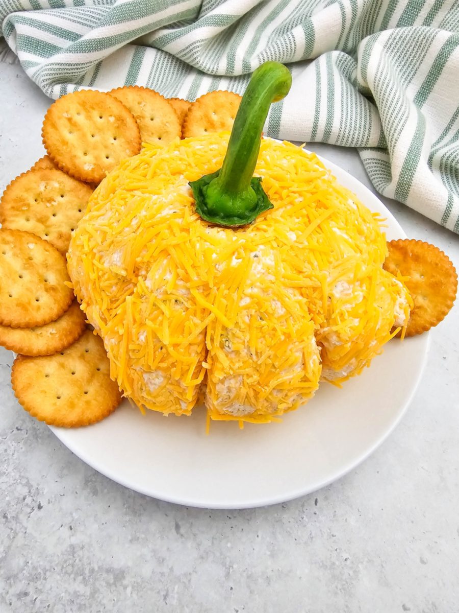 A slightly elevated, angled view of the complete ranch flavored cheese ball on a white plate. It is covered in shredded orange cheddar cheese, shaped like a pumpkin, and surrounded by crackers.
