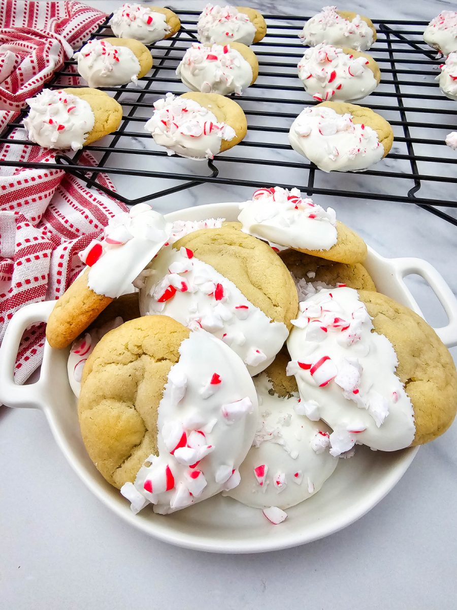 Christmas cookies piled high in a white serving dish, with more cookies cooling on a wire rack behind them.