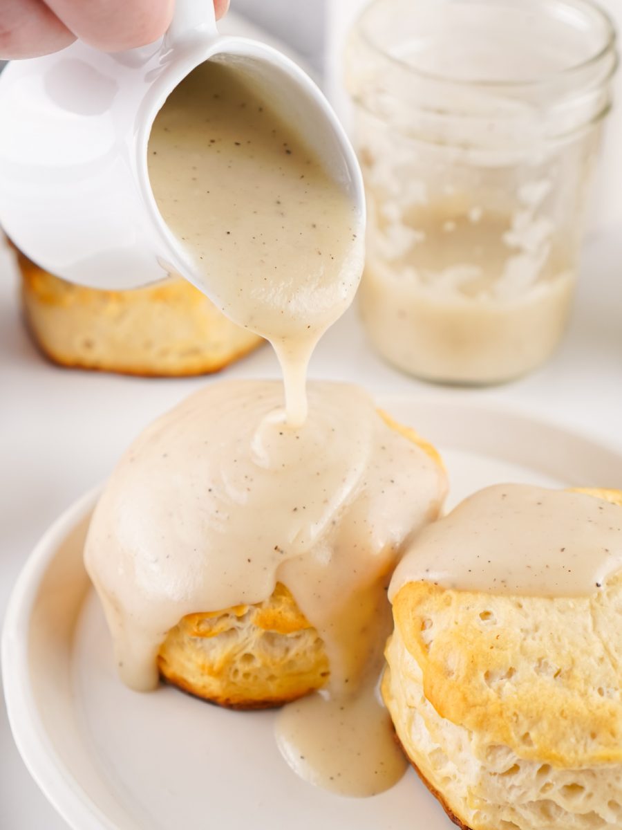 A hand pouring thick, creamy white country gravy from a white pitcher generously over two golden biscuits on a white plate.
