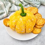 An angled, eye-level view of the Pumpkin Cheese Ball on a white plate, coated in shredded orange cheddar and topped with a green bell pepper stem. Round butter crackers are arranged around the cheese ball, with a folded green and white striped towel in the background.
