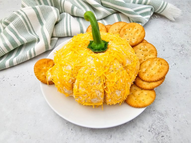 An angled, eye-level view of the Pumpkin Cheese Ball on a white plate, coated in shredded orange cheddar and topped with a green bell pepper stem. Round butter crackers are arranged around the cheese ball, with a folded green and white striped towel in the background.