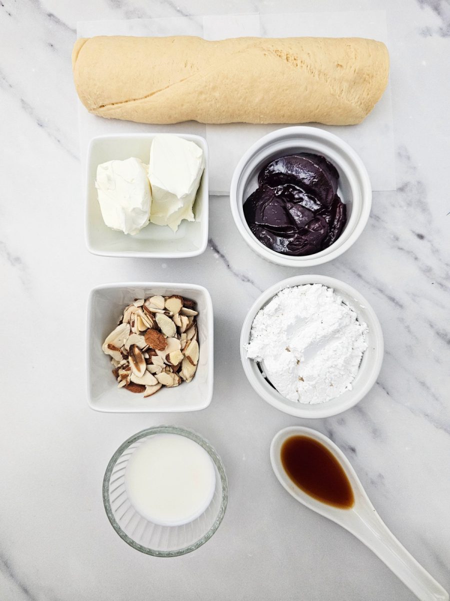 Overhead shot of all the ingredients: a crescent roll dough log, cream cheese, raspberry jam, powdered sugar, sliced almonds, milk, and vanilla extract, arranged in small white bowls on a marble background.