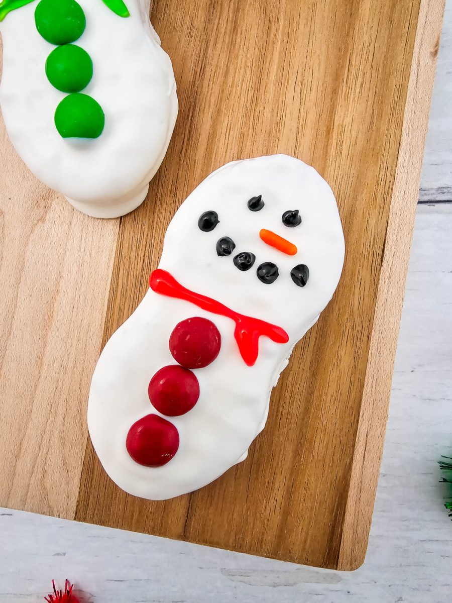 Close-up shot of a single Nutter Butter Snowman cookie decorated with a bright red gel scarf and red M&M buttons resting on a wooden cutting board.