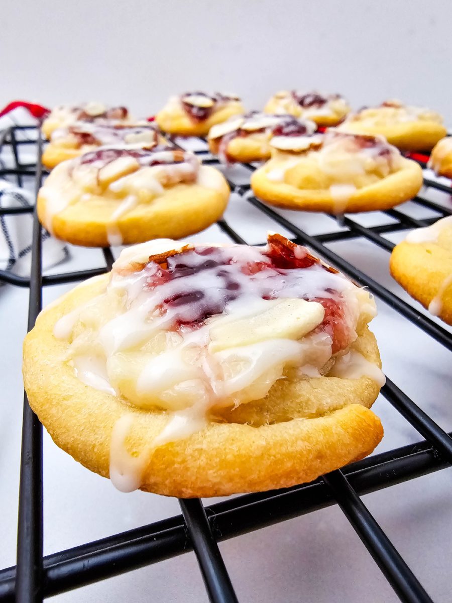Close up, eye-level view of a glazed raspberry cream cheese crescent cookie resting on a black wire cooling rack, with other cookies blurred in the background.