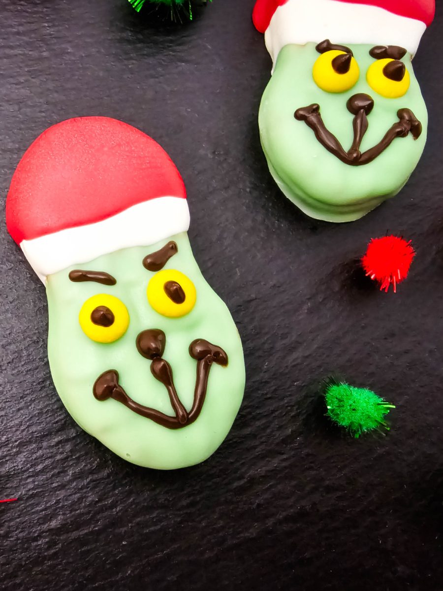 A very close-up detail shot of two finished Grinch Nutter Butter Cookies on a black slate, highlighting the smile, eyes, and the red and white Santa hat detail.