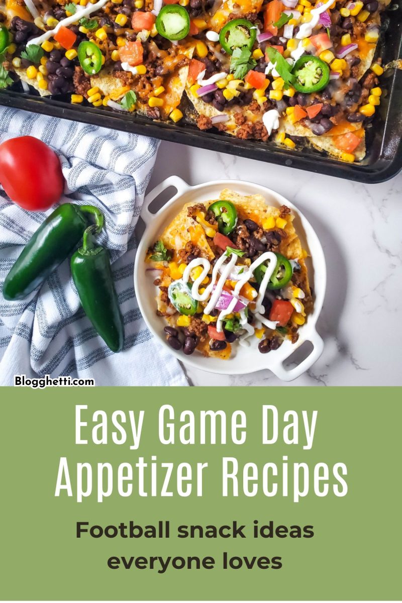 A close-up overhead view of a large sheet pan of loaded beef nachos and a smaller white bowl containing a single serving. The nachos are topped with seasoned ground beef, black beans, melted cheese, fresh jalapeño slices, tomatoes, and a drizzle of sour cream. Two whole jalapeños and a striped kitchen towel sit on the marble countertop next to the bowl.