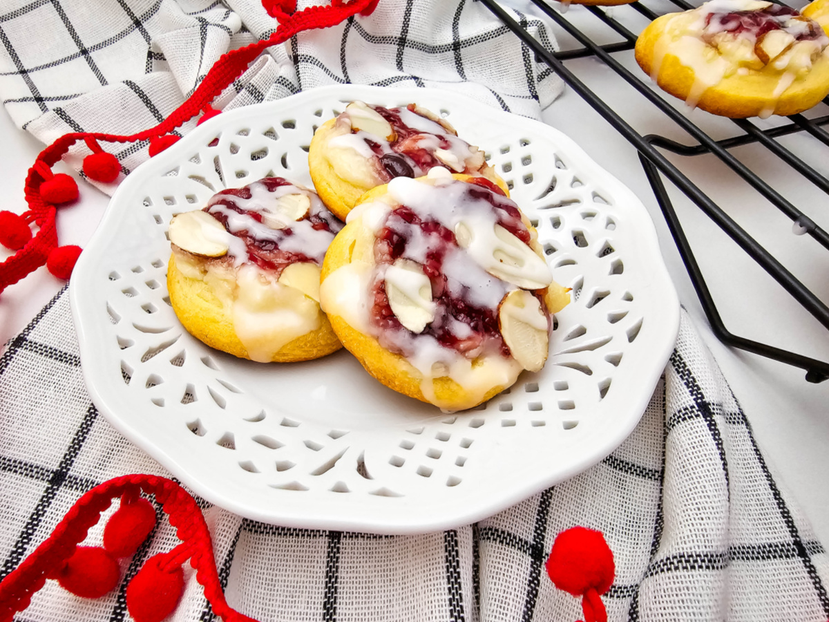 Three glazed raspberry cream cheese crescent cookies on a white decorative plate, with a black and white checkered cloth and red pom-pom trim. A black cooling rack is visible in the background.