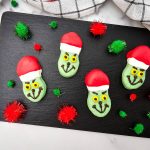 Four completed Grinch Nutter Butter Cookies with red and white Santa hats and dark chocolate facial details, arranged on a black slate serving tray with red and green pom-poms.