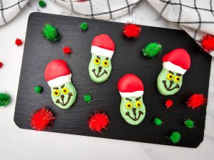 Four completed Grinch Nutter Butter Cookies with red and white Santa hats and dark chocolate facial details, arranged on a black slate serving tray with red and green pom-poms.