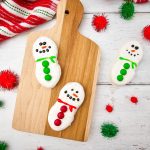 Feature image showing three decorated Nutter Butter Snowman Cookies (two with green scarves/buttons, one with red) arranged on a wooden cutting board on a white wood surface with holiday towel and red and green pom-poms.