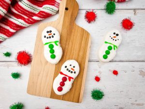 Feature image showing three decorated Nutter Butter Snowman Cookies (two with green scarves/buttons, one with red) arranged on a wooden cutting board on a white wood surface with holiday towel and red and green pom-poms.