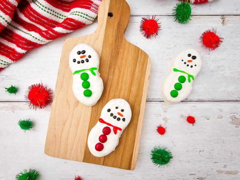 Feature image showing three decorated Nutter Butter Snowman Cookies (two with green scarves/buttons, one with red) arranged on a wooden cutting board on a white wood surface with holiday towel and red and green pom-poms.
