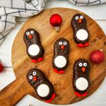 Top-down view of six finished chocolate-coated peanut butter cookies decorated like penguins, centered on a wooden cutting board with a festive napkin and red ornaments.