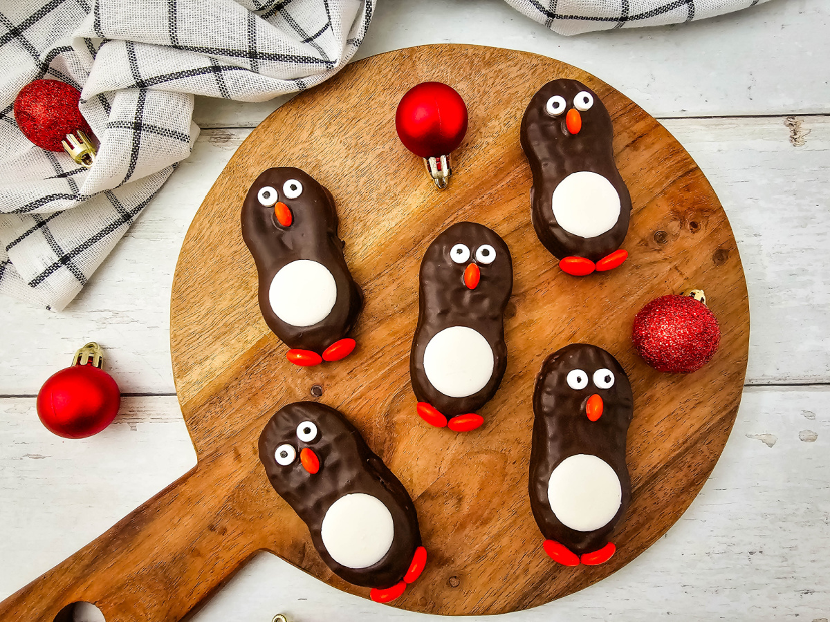 Top-down view of six finished chocolate-coated peanut butter cookies decorated like penguins, centered on a wooden cutting board with a festive napkin and red ornaments.