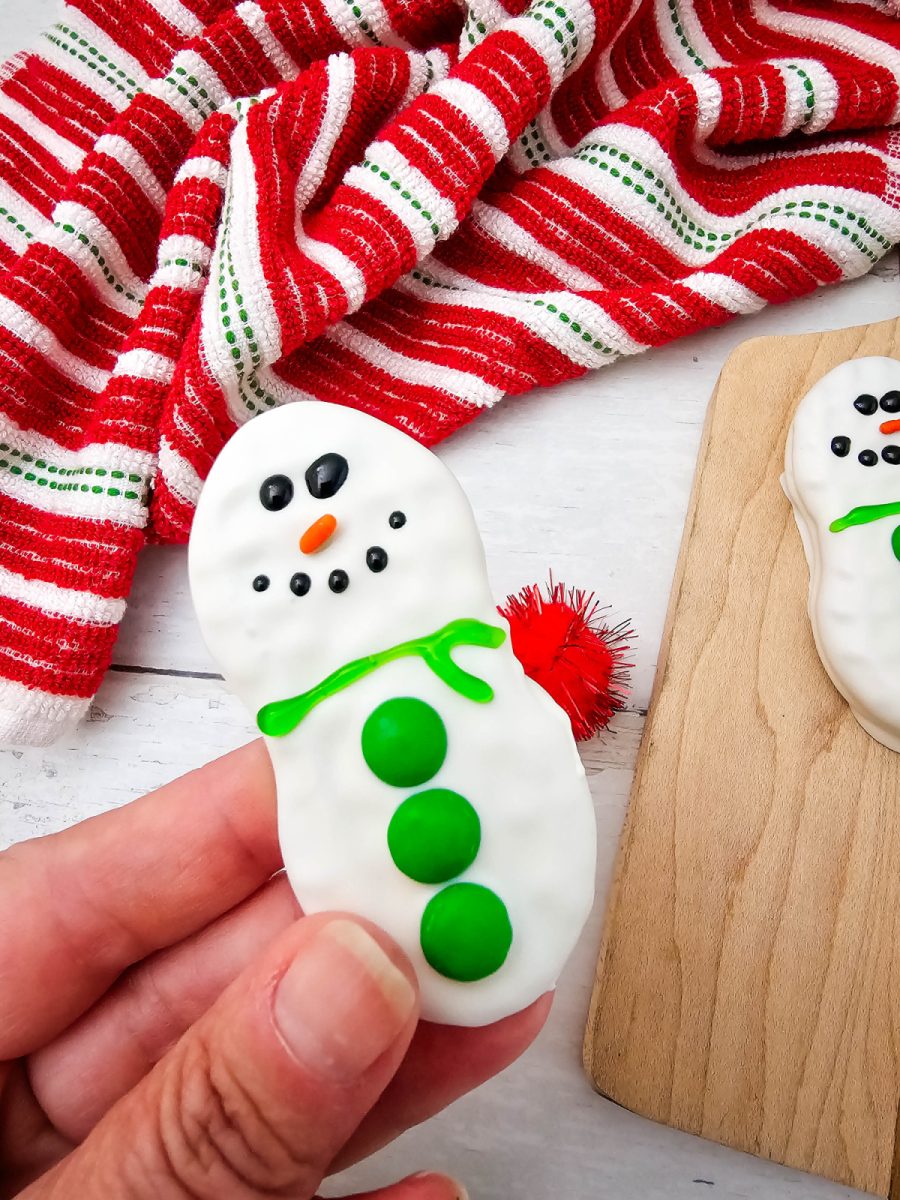 A hand holding a single Nutter Butter Snowman cookie decorated with white coating, a bright green gel scarf, green M&M buttons, black eyes, and an orange sprinkle nose, against a white wooden surface and red and white striped fabric.
