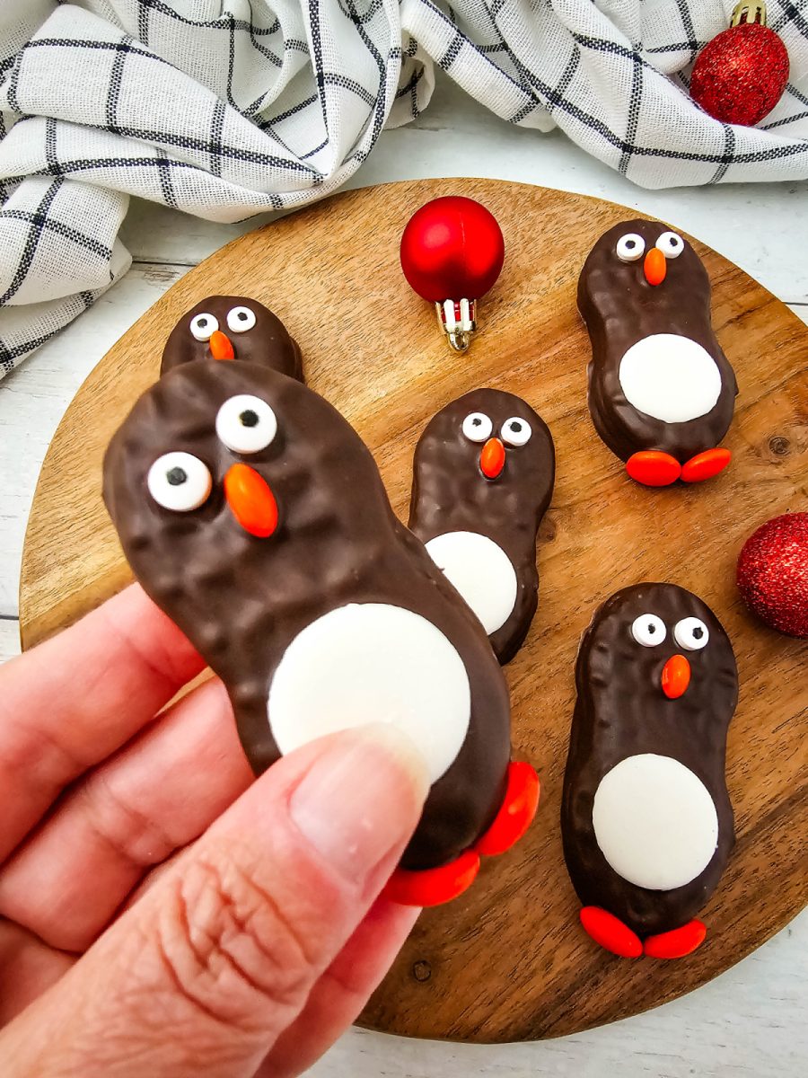 A hand holding one finished chocolate holiday treat with white tummy, candy eyes, and M&M feet, displayed against a wooden board and festive decor.