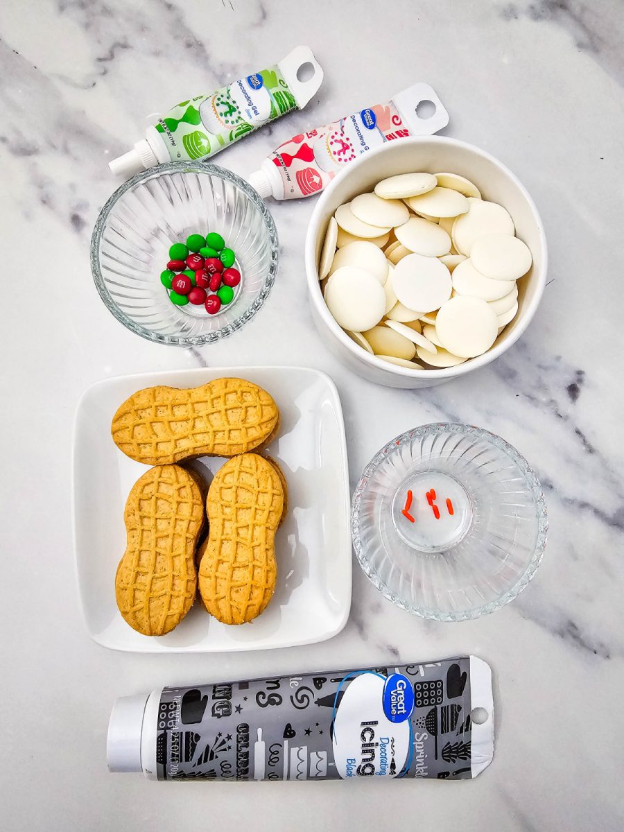 Overhead flat lay of all the ingredients needed for the recipe, including Nutter Butter cookies, white candy melts, red and green M&Ms, red and green decorating gel tubes, black decorating icing, and orange sprinkles.