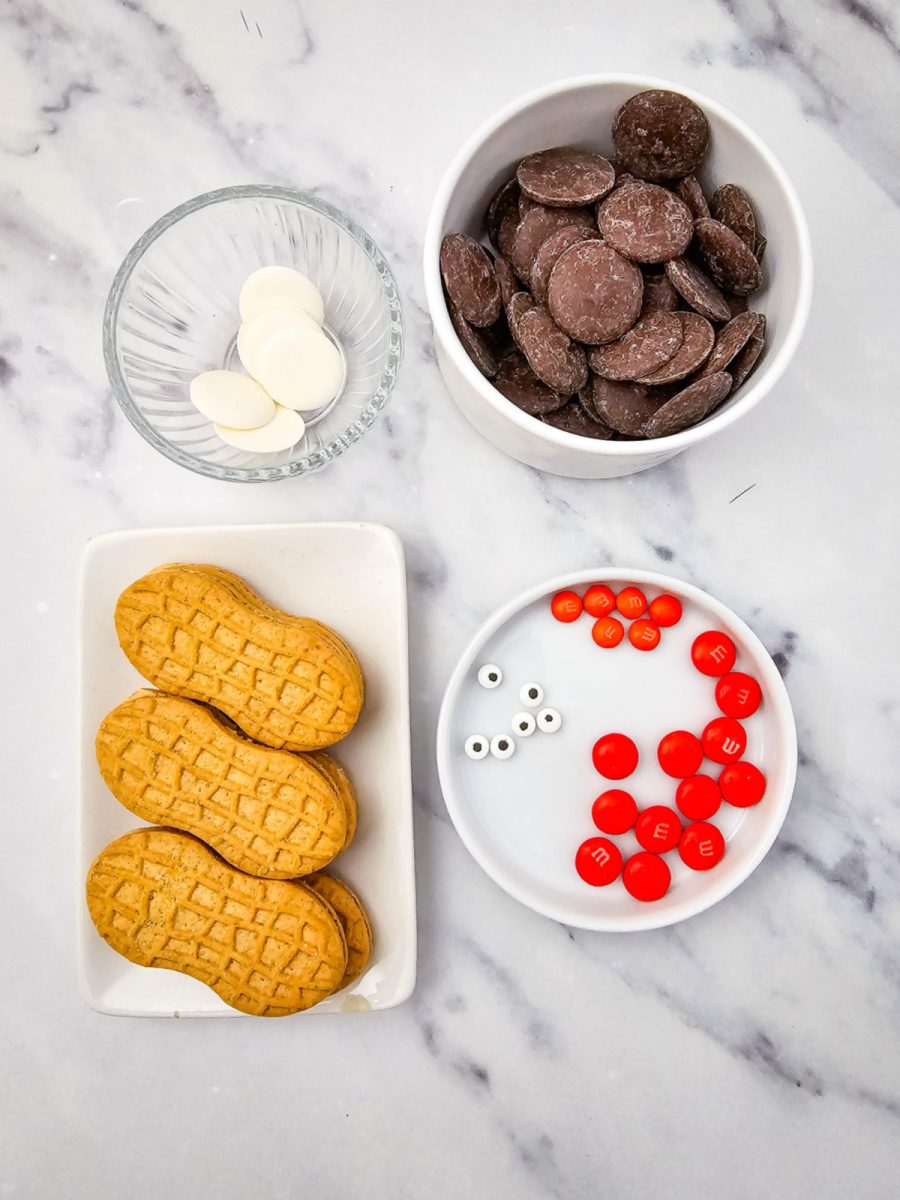 A flat lay of all the simple ingredients: sandwich cookies, dark chocolate wafers, white candy melts, candy eyeballs, and orange M&Ms, arranged on a marble background.