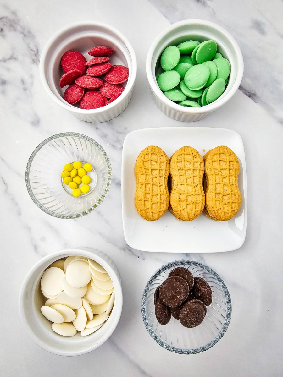 An overhead flat lay of all the Grinch cookie ingredients: bowls of red, green, white, and dark chocolate candy melts, yellow M&Ms, and three Nutter Butter cookies on a marble surface.