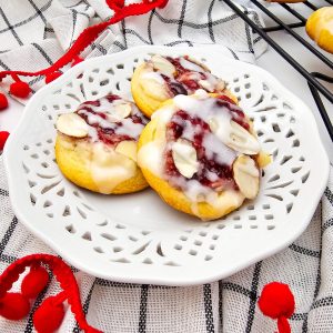 Three finished raspberry cream cheese crescent cookies on a white decorative plate, drizzled with glaze and topped with sliced almonds.