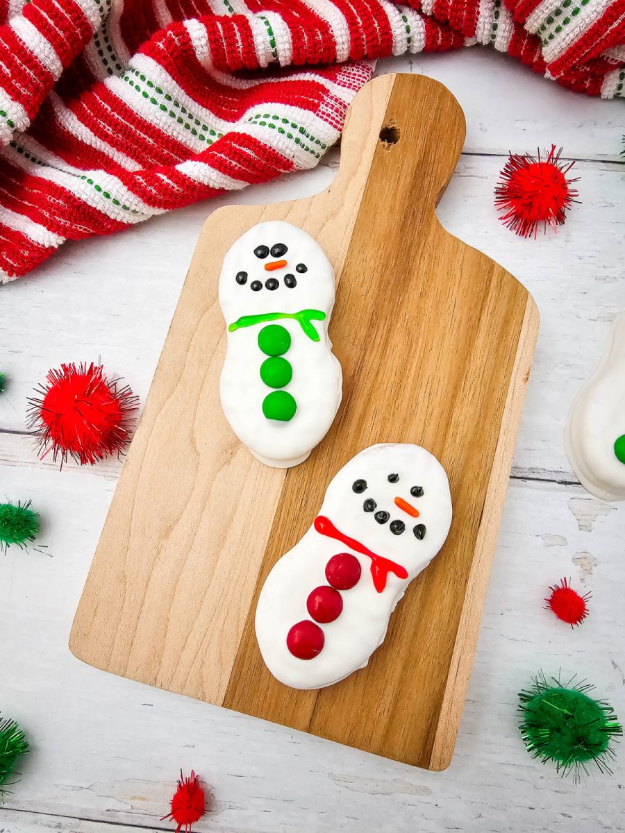Overhead flat lay showing two decorated Nutter Butter Snowman Cookies on a wooden cutting board, one with a green scarf/buttons and one with a red scarf/buttons, with a red and white striped towel and festive pom-poms scattered around.