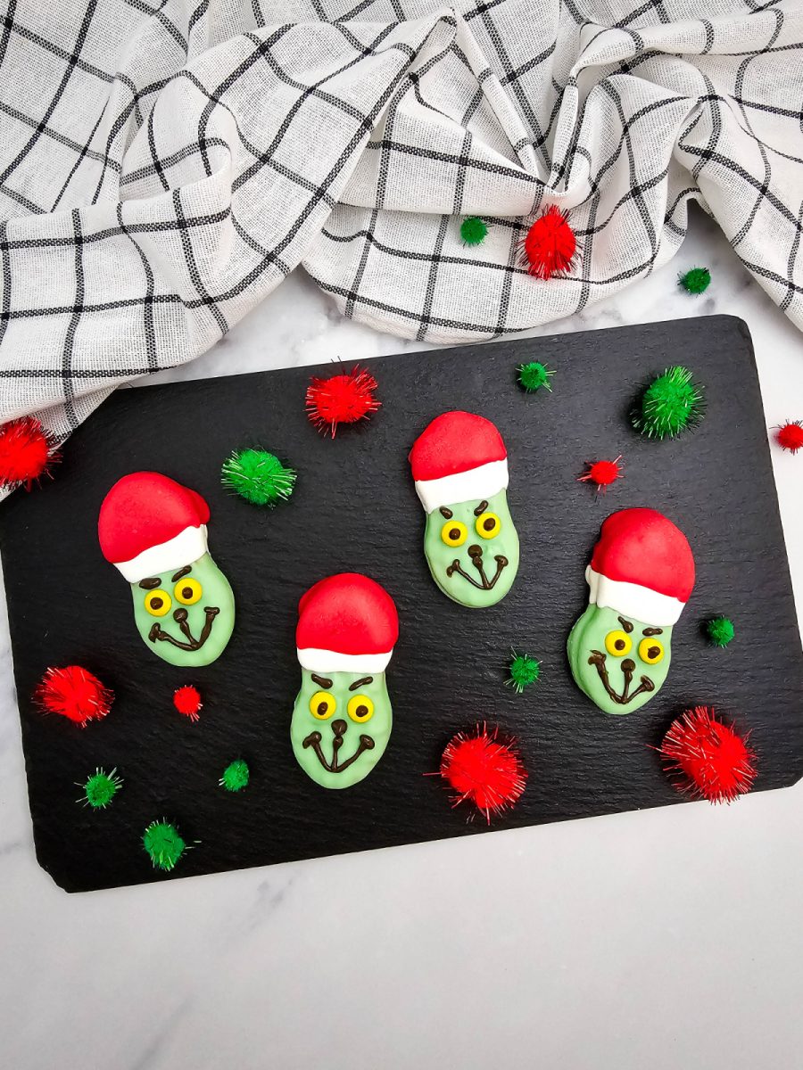 Overhead view of four finished Grinch Nutter Butter Cookies set on a black slate tray, surrounded by small red and green tinsel decorations and a white checkered cloth.