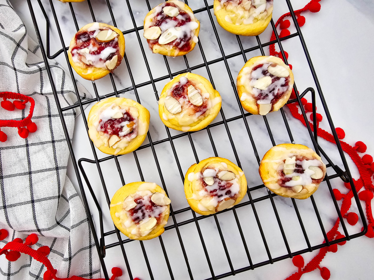 Overhead view of nine glazed raspberry cream cheese crescent cookies cooling on a black wire rack, with red pom-pom trim and a checkered cloth on the white marble surface.
