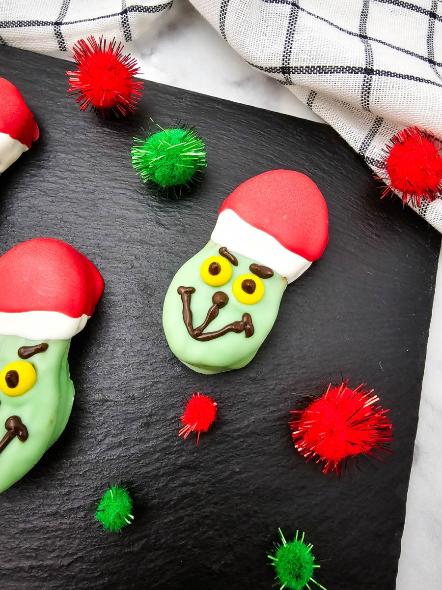 A close-up shot of a single completed Grinch Nutter Butter Cookie on a black slate surface, showing the green face, red hat, yellow M&M eyes, and dark chocolate smile and eyebrows.