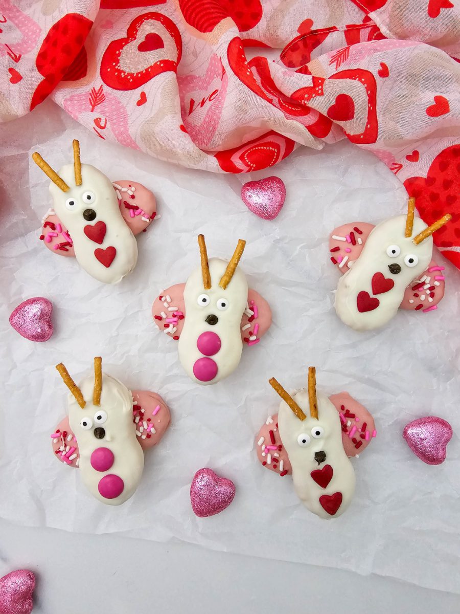 Six Nutter Butter love bug cookies arranged on white parchment paper decorated with pink glitter hearts and a red heart-patterned fabric background.