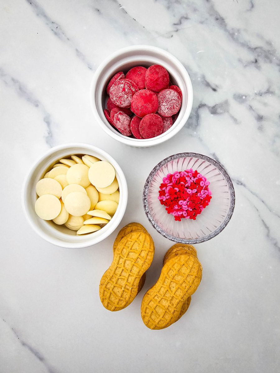 A top-down view of the ingredients needed for the recipe, including white chocolate melting wafers, red candy melts, festive red and pink sprinkles in a glass bowl, and several peanut-shaped sandwich cookies on a marble surface.