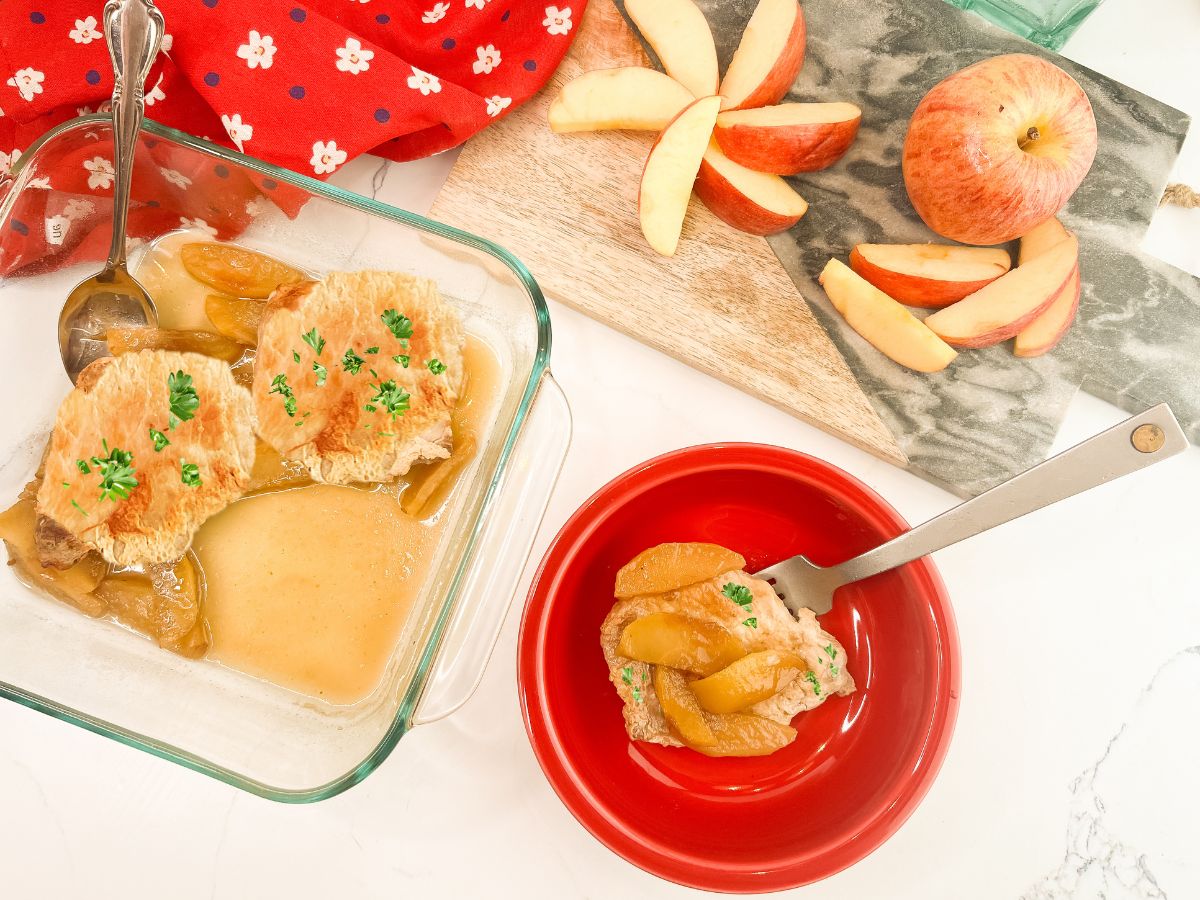 A wide-angle tabletop scene. On the left is a glass dish with two pork chops in a light sauce; on the right is a red bowl containing a single serving. In the background, fresh red apple slices are arranged in a fan shape on a wooden and marble cutting board.
