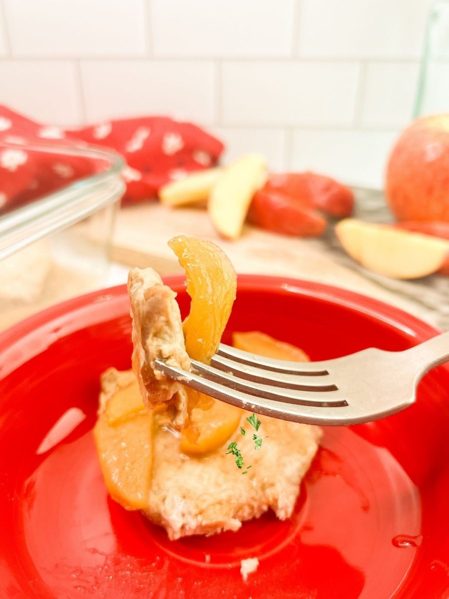 A macro shot focusing on a silver fork held in the air. On the tines of the fork is a small, square bite of cooked pork topped with a glistening, tender slice of apple. The blurred red bowl and white tile backsplash are in the background.