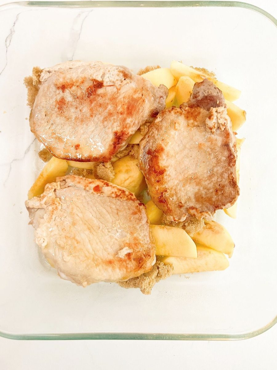 A top-down view of three thick, boneless pork chops that have been lightly seared golden-brown on the surface. They are layered over a bed of pale yellow sliced apples and mounds of crumbly light brown sugar in a clear square glass baking dish.