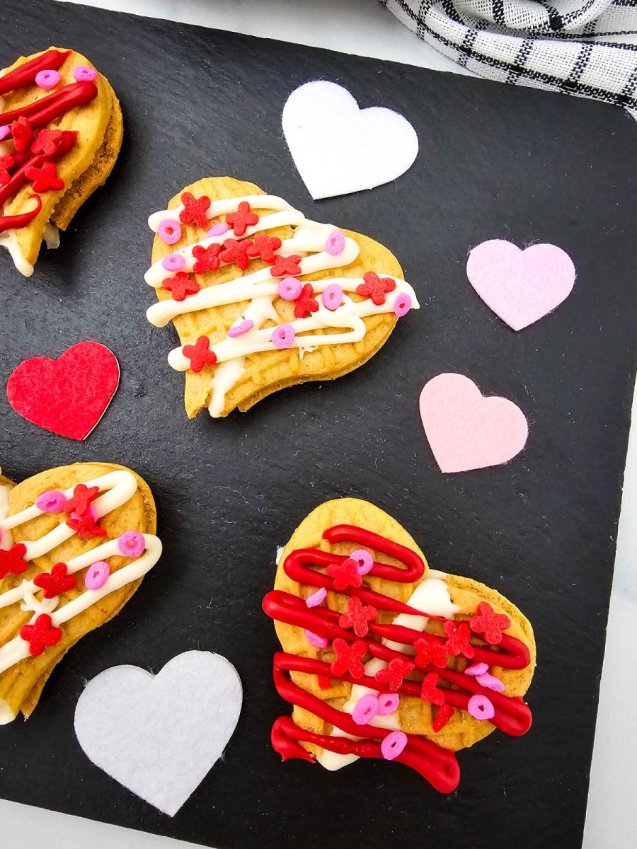 A detailed vertical shot highlighting the texture of the no-bake peanut butter treats. The cookies feature a distinct waffle pattern and are heavily decorated with colorful icing and sprinkles. Small felt heart cutouts in pink, red, and white are scattered around the treats on a dark slate tray.