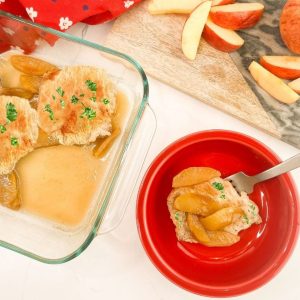 A square-cropped image showing a serving of pork and apples in a red bowl in the foreground. In the upper left, the remaining pork chops are visible in the glass baking pan, and fresh apple slices rest on a wooden board in the background.