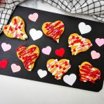 A wide, bright feature shot of six decorated Nutter Butter heart cookies on a black rectangular serving board. The composition includes a stylish checkered kitchen towel in the corner and a variety of colorful heart shapes, making it a festive presentation for Valentine's Day.