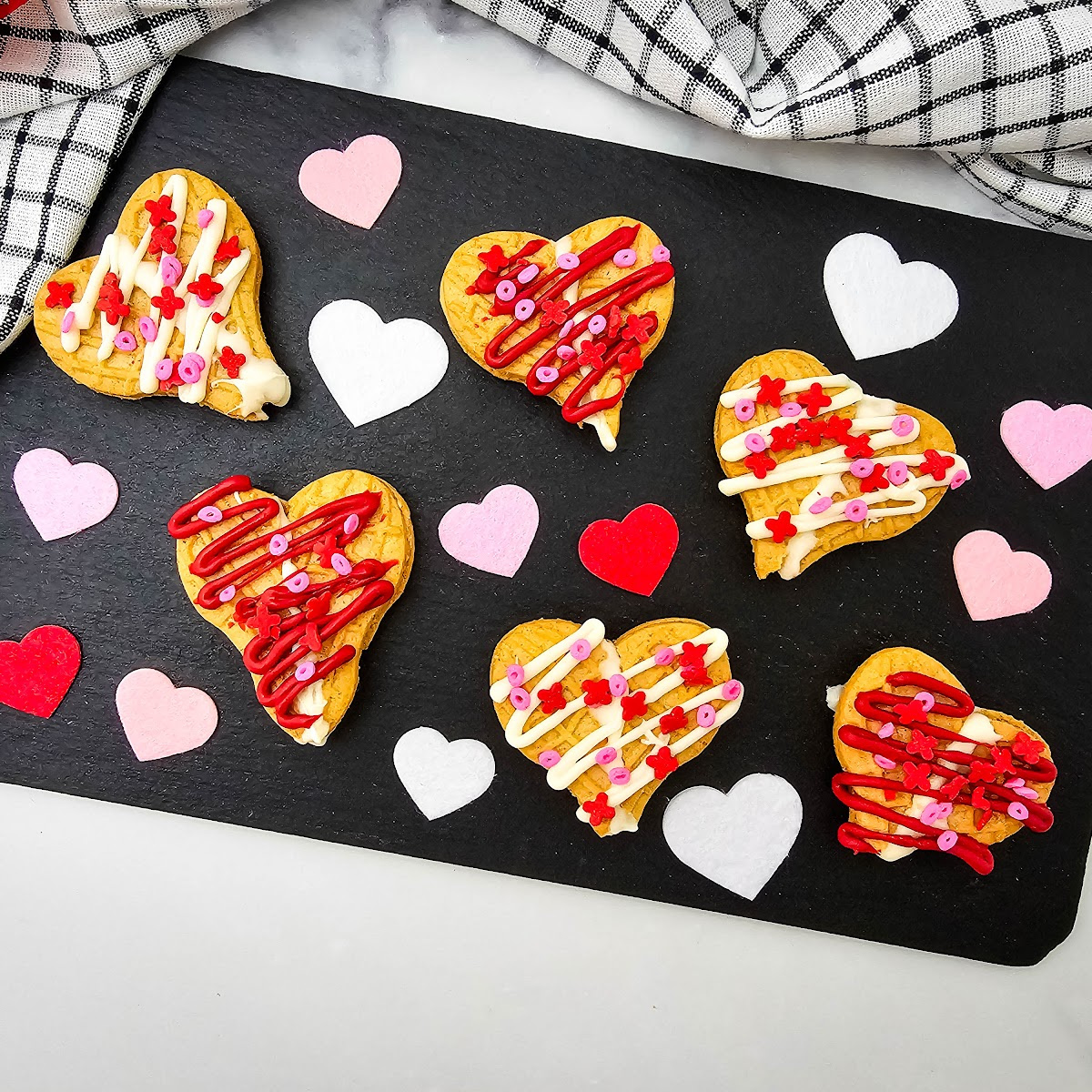 A wide, bright feature shot of six decorated Nutter Butter heart cookies on a black rectangular serving board. The composition includes a stylish checkered kitchen towel in the corner and a variety of colorful heart shapes, making it a festive presentation for Valentine's Day.