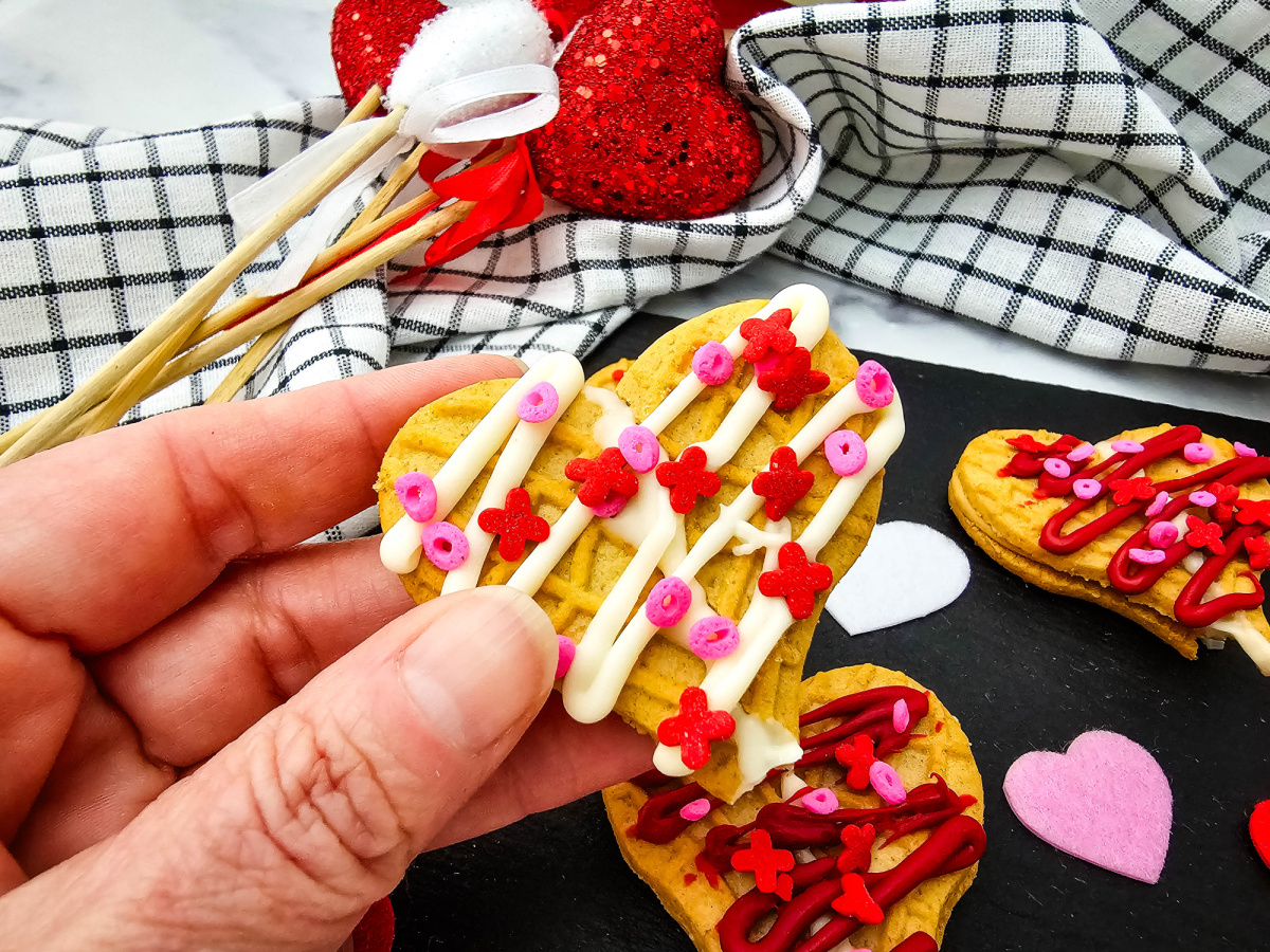 A close-up shot of a hand holding one decorated heart-shaped peanut butter cookie. The cookie is drizzled with white chocolate and adorned with red and pink sprinkles. In the background, sparkly red heart picks and a checkered napkin create a warm, celebratory atmosphere.