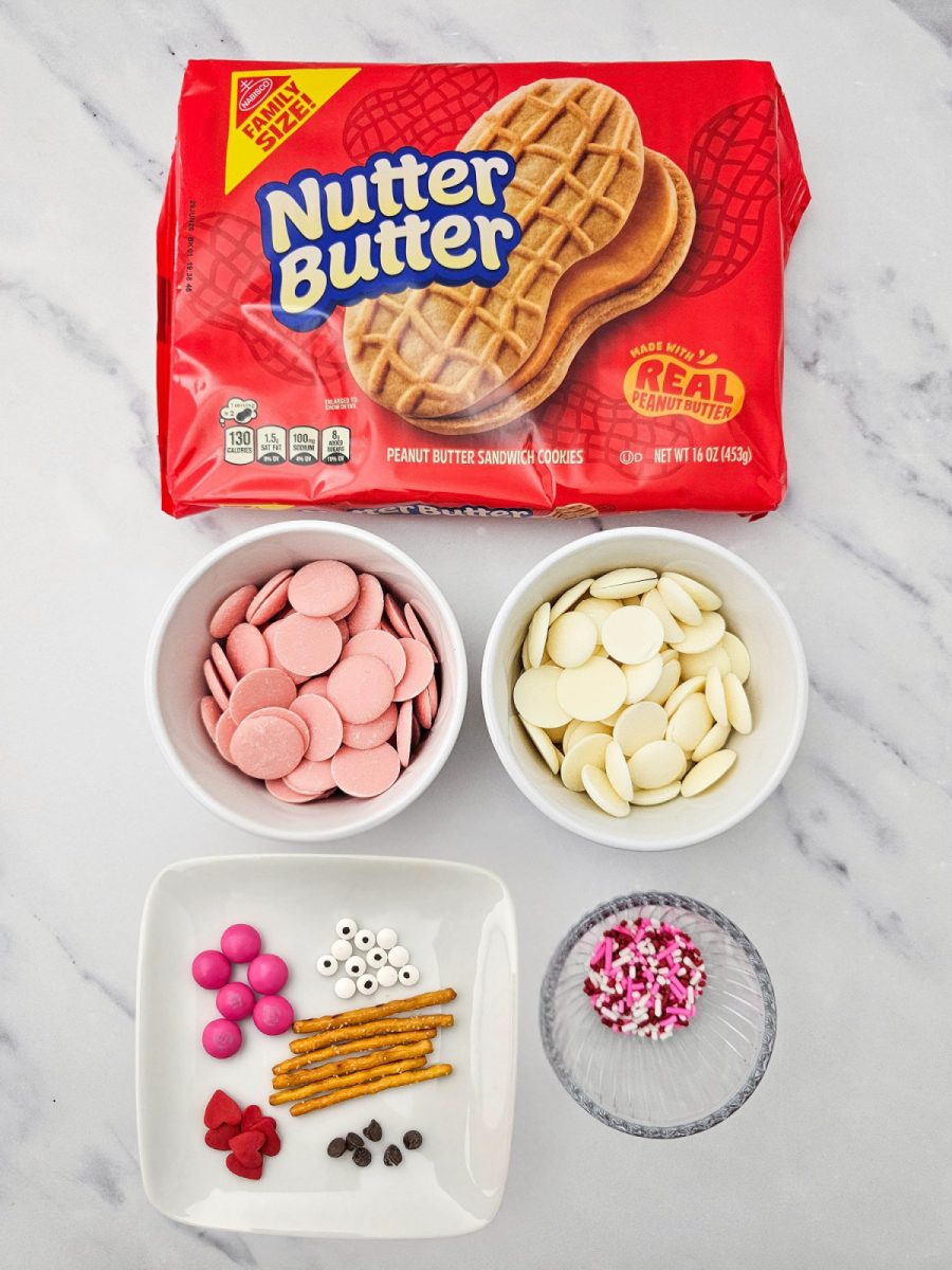 An overhead shot of the recipe ingredients including a package of Nutter Butters, bowls of pink and white candy melts, pretzels, sprinkles, and candy eyes.