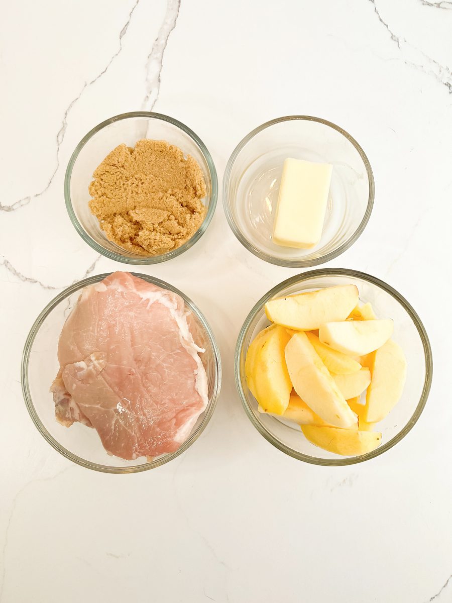 An overhead shot of four small glass bowls arranged on a white marble surface. The bowls contain raw pink boneless pork chops, a pile of sliced peeled apples, a heap of moist light brown sugar, and a rectangular pat of yellow butter.