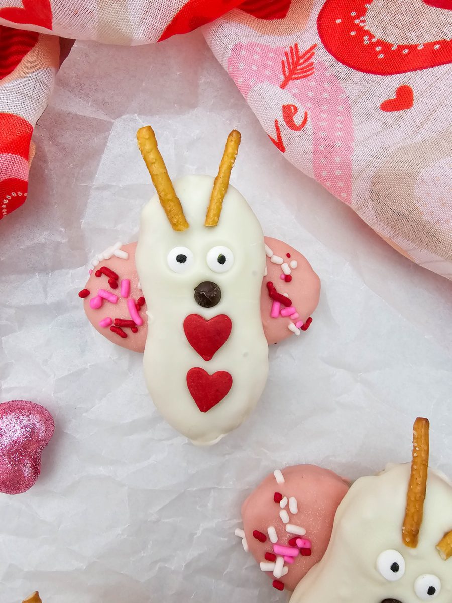 A close-up of a love bug cookie decorated with two red heart sprinkles on its body, resting on white parchment paper next to a pink glitter heart.