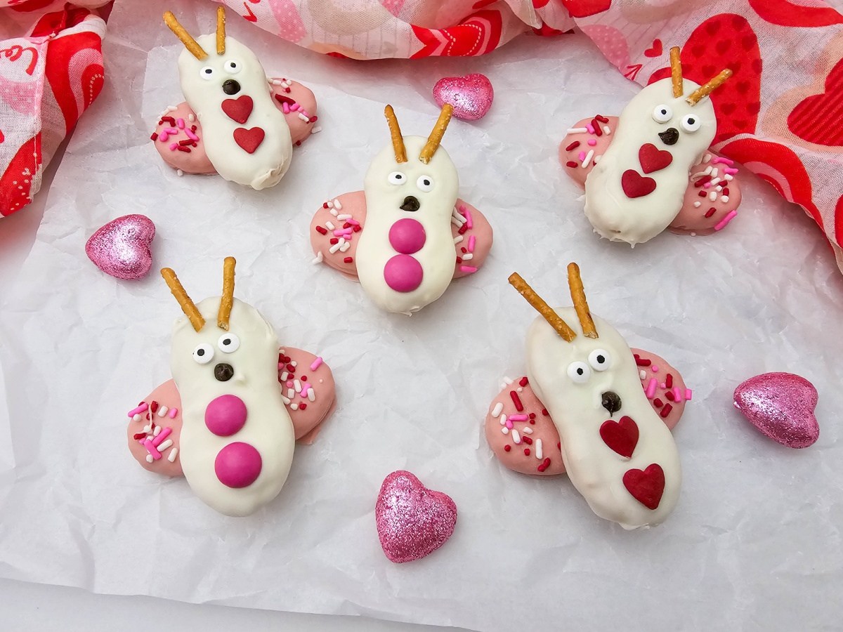 A wide overhead shot of six Nutter Butter love bug cookies spread across parchment paper with scattered pink hearts and a red scarf.