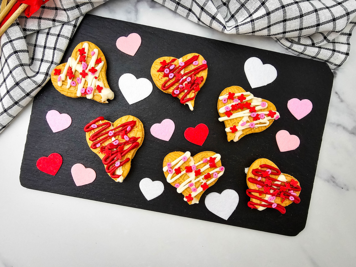 An overhead shot of six heart-shaped sandwich cookies decorated with red and white chocolate drizzles and colorful sprinkles. The cookies are arranged on a black rectangular board alongside scattered pink, white, and red felt hearts, set against a marble background with a checkered napkin.