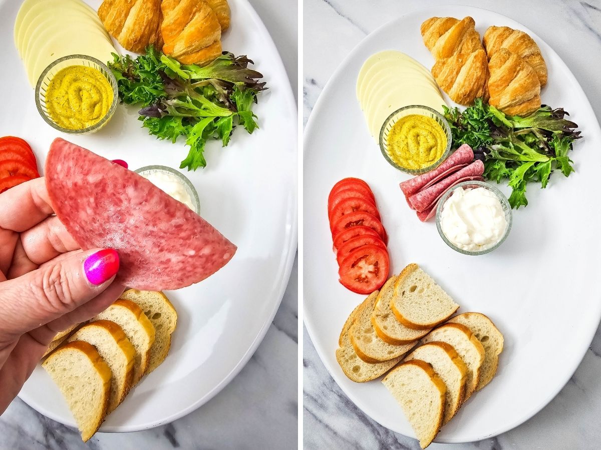 A process photo demonstrating how to build a deli spread. A hand with pink nail polish holds a slice of hard salami over a tray already featuring assorted cheeses, bread slices, fresh lettuce, and dipping sauces.