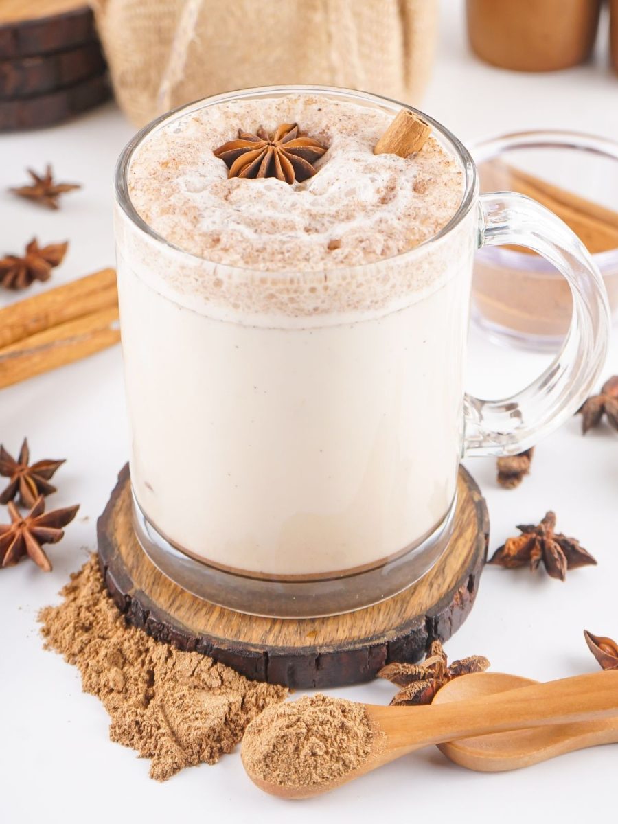 A frothy tea latte in a clear glass mug topped with a star anise and cinnamon stick sitting on a rustic wooden coaster.